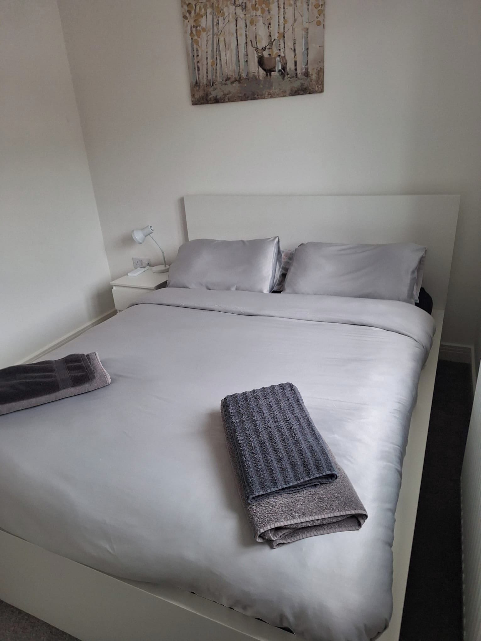A neatly made bed with pillows and a folded towel, a bedside lamp, and a wall-mounted painting of a forest scene.