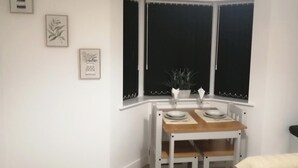 Una habitación de esquina con una mesa y sillas blancas, una ventana con cortinas negras y tres cuadros en la pared.