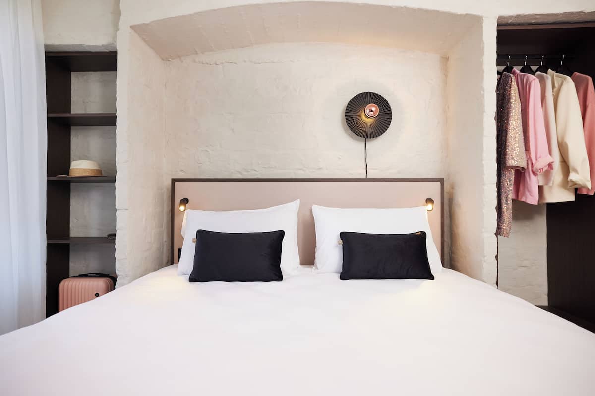 A neatly made bed with two black pillows, a round wall clock, and a clothing rack with various garments.