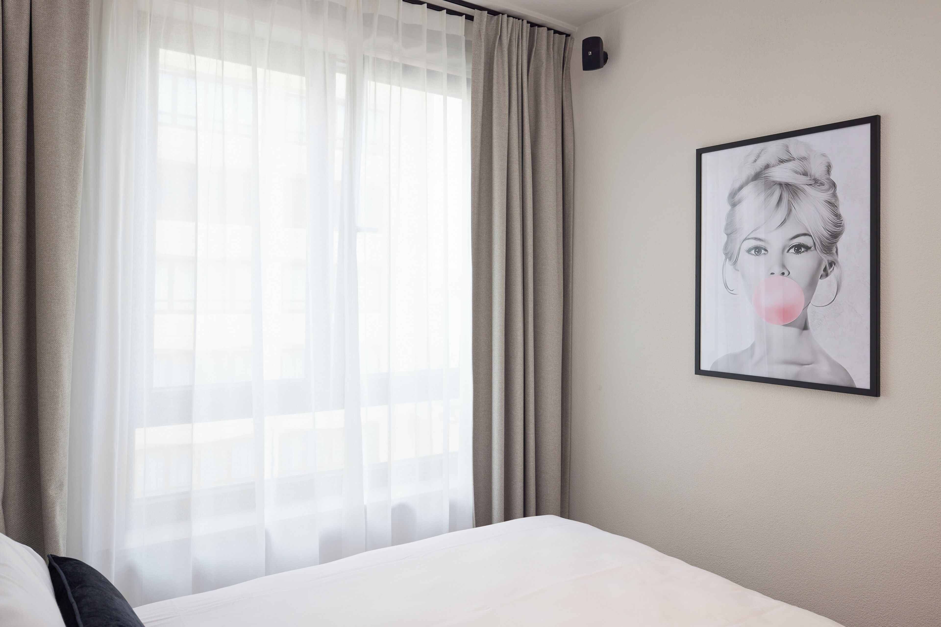 Quarto de hotel com uma cama, uma janela com cortinas translúcidas e uma obra de arte emoldurada na parede.