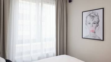 Quarto de hotel com uma cama, uma janela com cortinas translúcidas e uma obra de arte emoldurada na parede.
