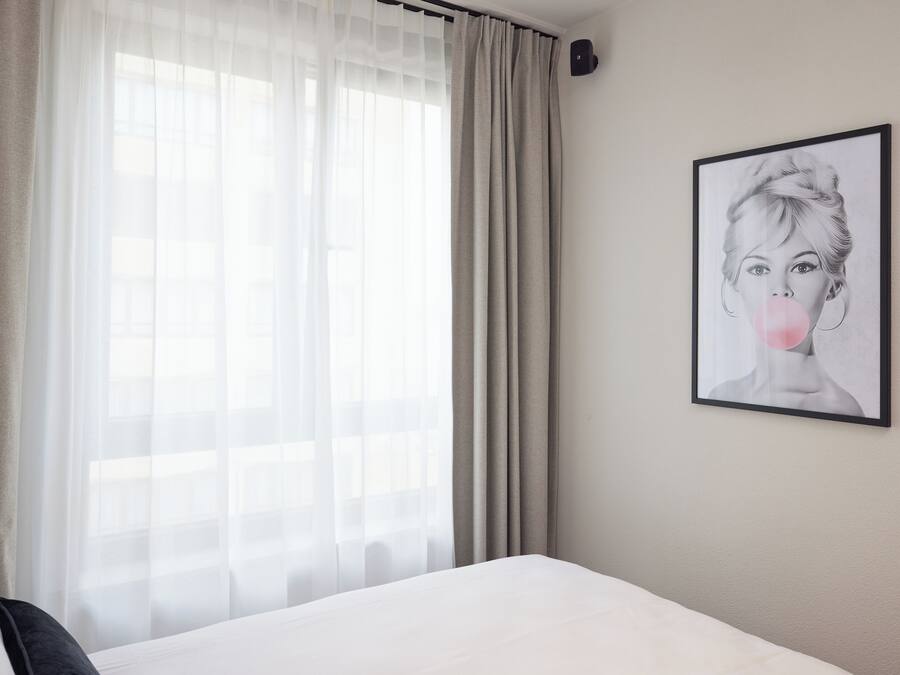 A hotel room with a bed, a window with sheer curtains, and a framed artwork on the wall.