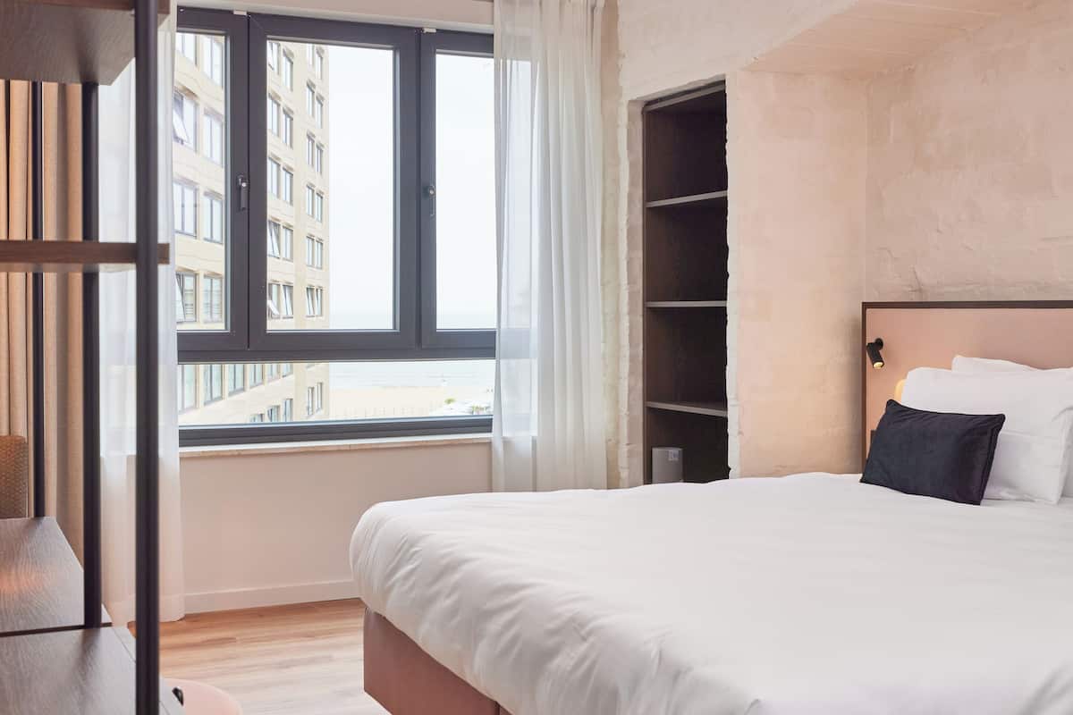 A hotel room with a large bed, a window with curtains, a wooden floor, and a view of a building outside.