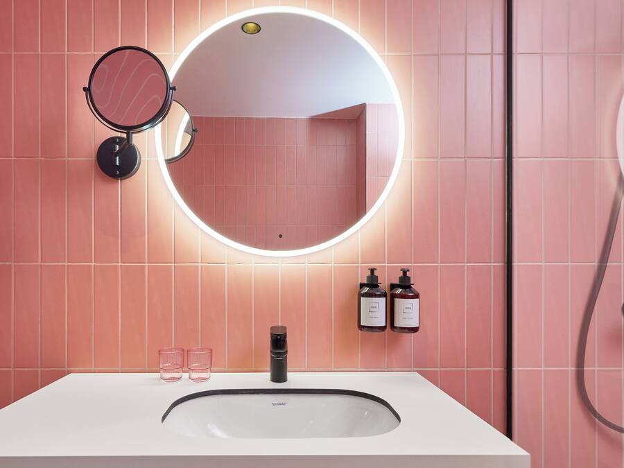 Een badkamer met een wit wastafel, twee ronde spiegels, een ronde lamp boven de spiegel en twee flessen shampoo en conditioner aan de muur.