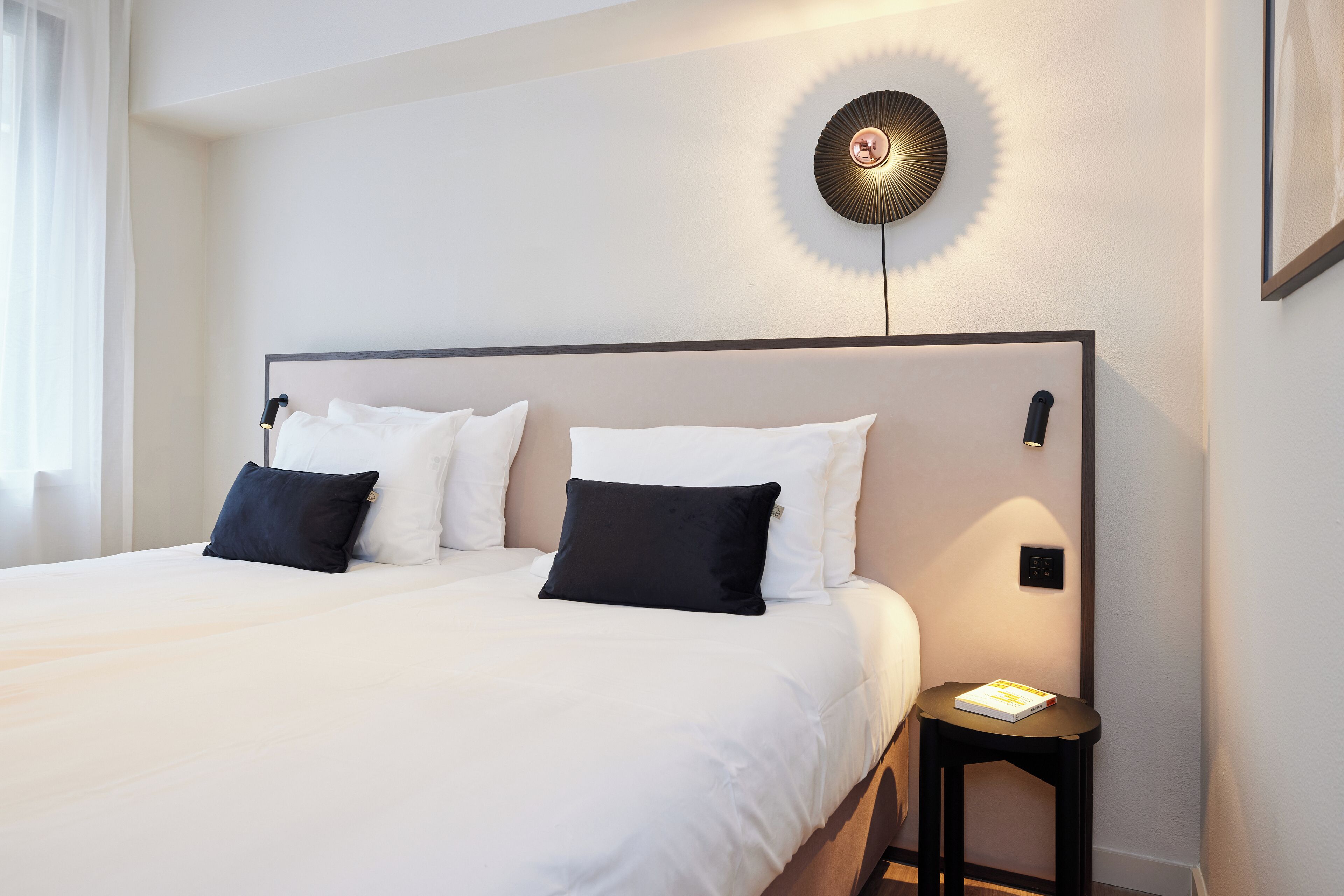 Una cama bien hecha con almohadas blancas y negras, una mesita de noche con un libro, y una lámpara de pared.