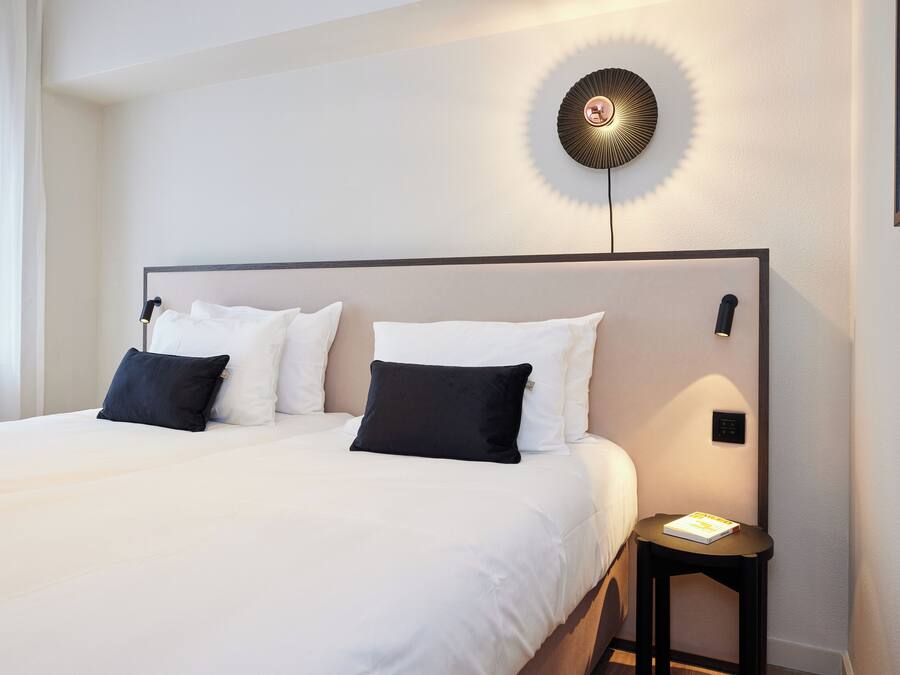 Una cama bien hecha con almohadas blancas y negras, una mesita de noche con un libro, y una lámpara de pared.