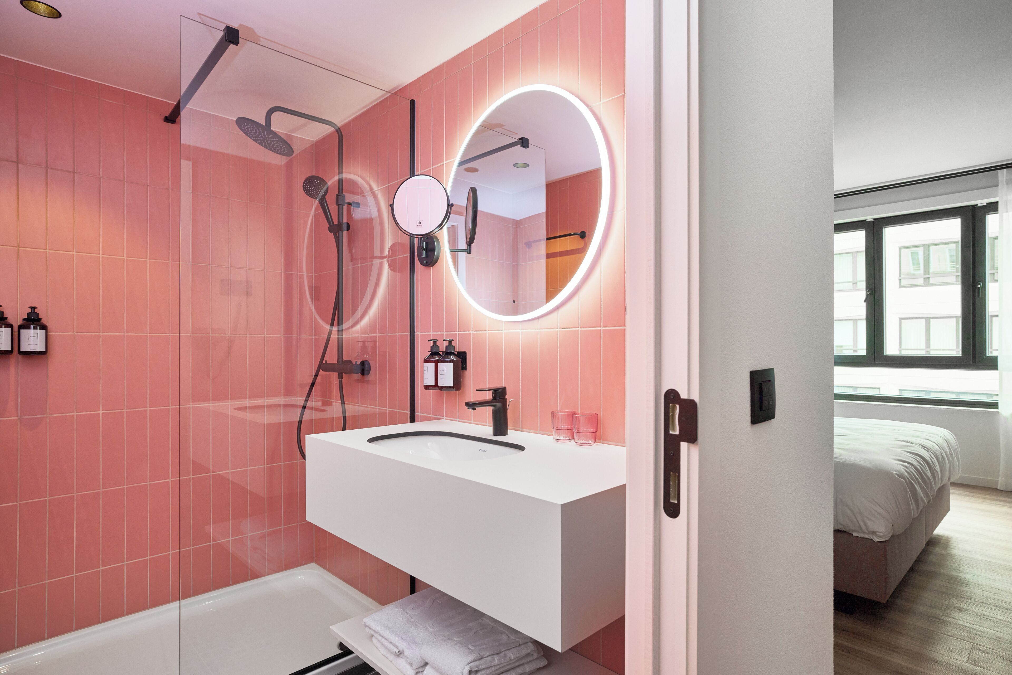 A modern bathroom with pink tiles, a white sink, a handheld showerhead, a round mirror with a light, and a visible window in the adjacent room.
