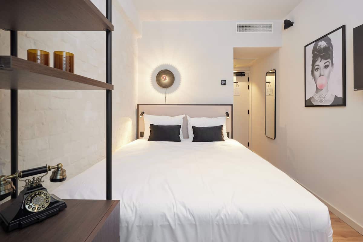 A modern hotel room with a large bed, a desk with a vintage telephone, a wall-mounted light, and a framed picture.
