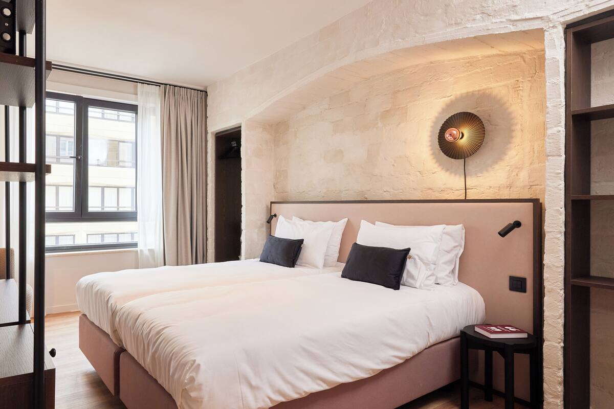 A bedroom with a large bed, a bedside table, a wall-mounted light, and a window with curtains.