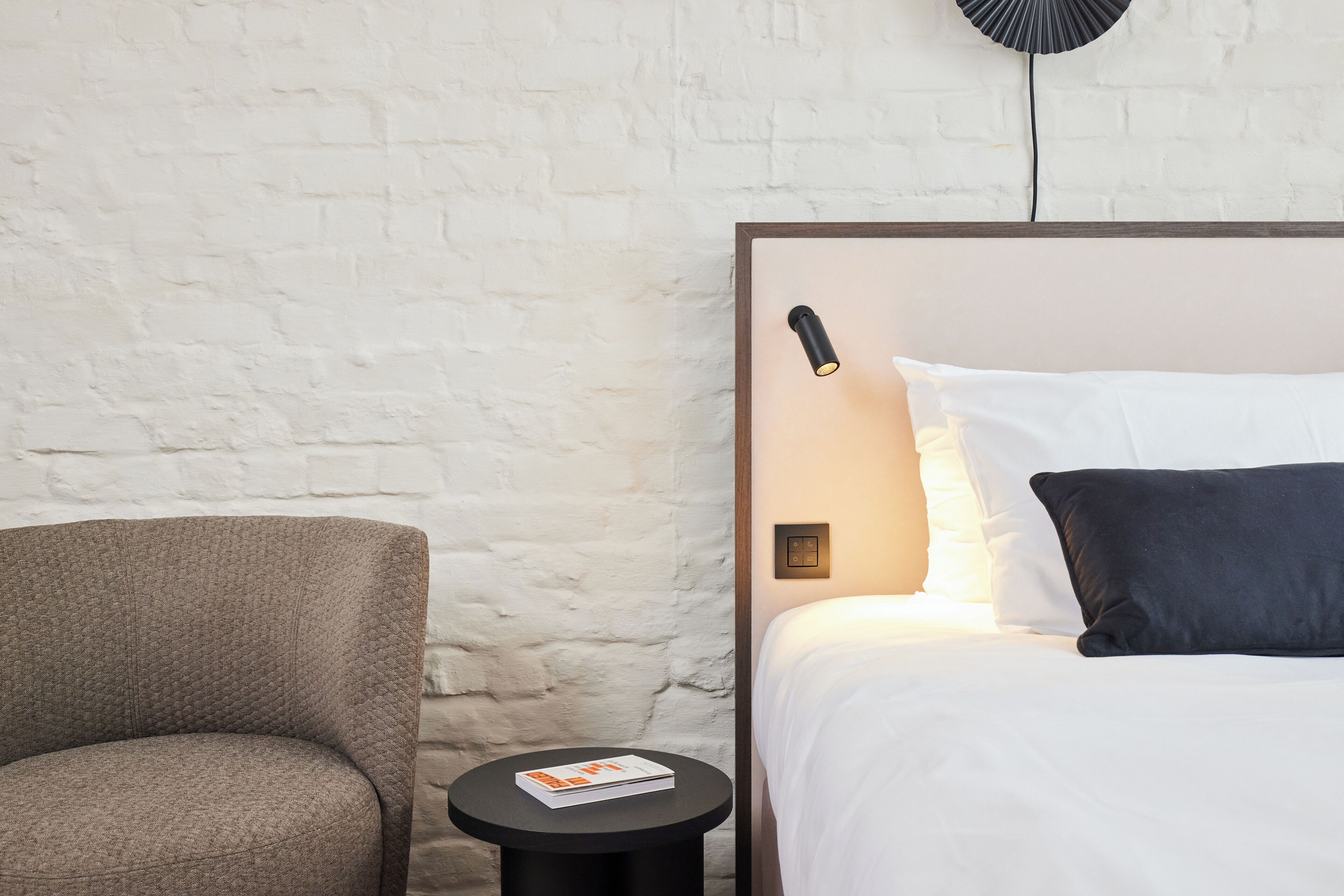 Una habitación de hotel con una cama, una mesita redonda, una silla y una pared de ladrillo blanco.
