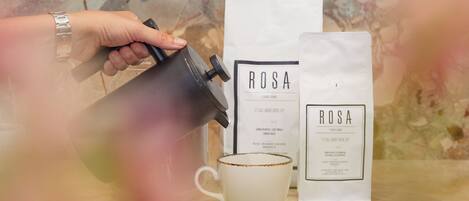 Una mano vertiendo café en una taza blanca con borde dorado, junto a dos bolsas blancas etiquetadas 'ROSA'.