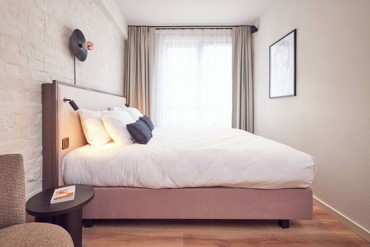 A modern hotel room with a large bed, a small side table, a wall-mounted fan, and a window with curtains.