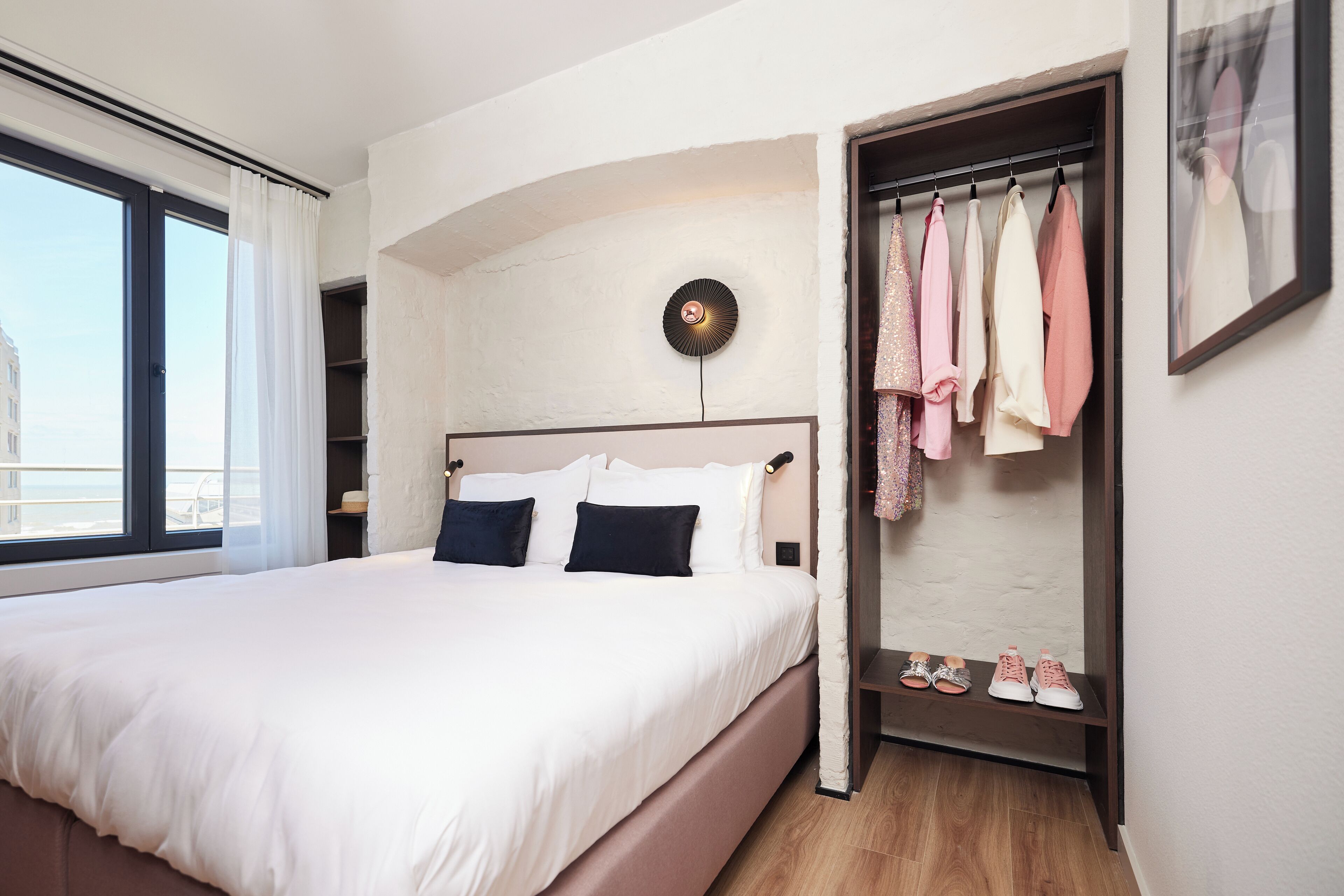 A hotel room with a large bed, a wardrobe with hanging clothes, and a view of the city through the window.