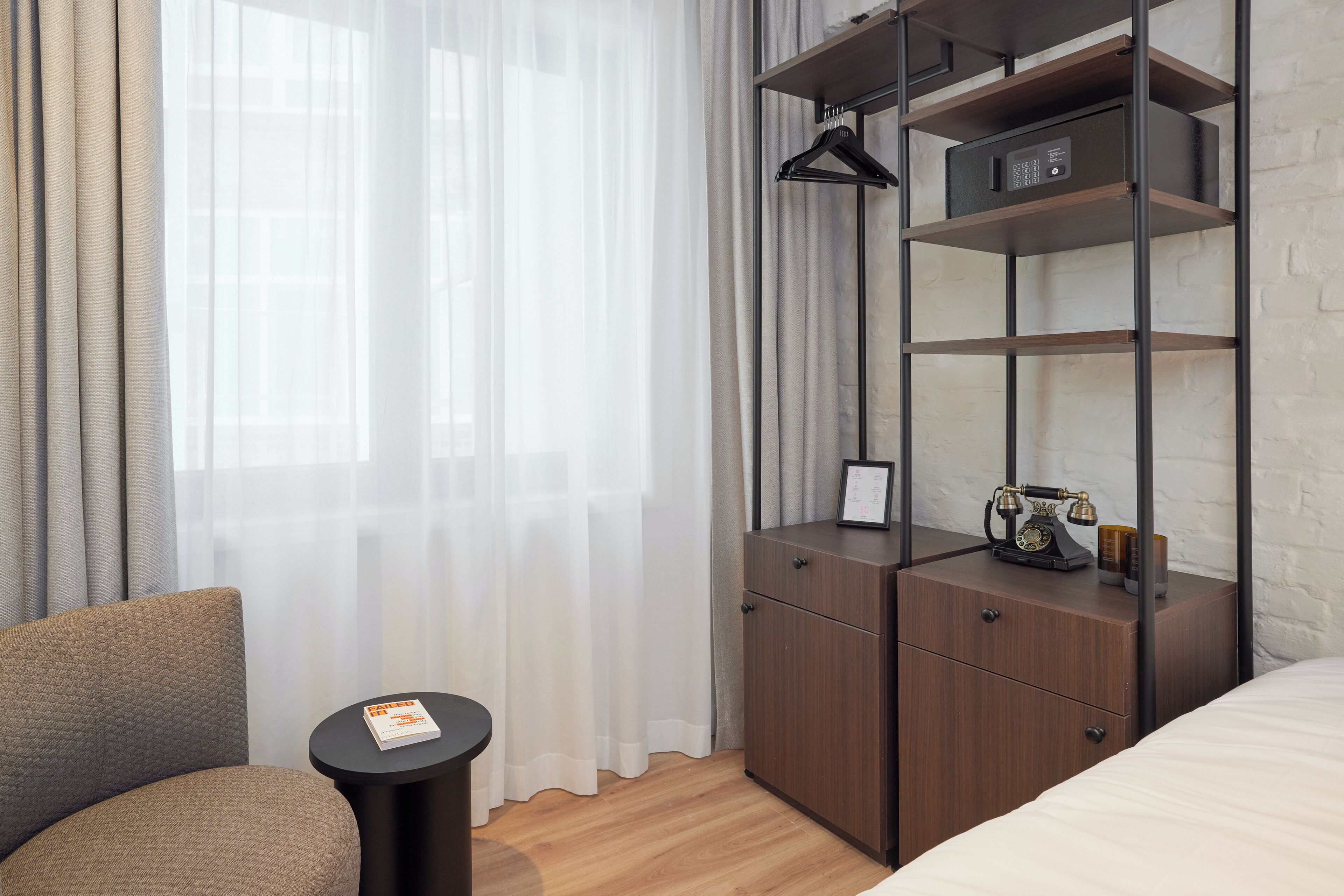 A hotel room with a bed, a wooden nightstand, a small table, a chair, and a wardrobe with shelves.