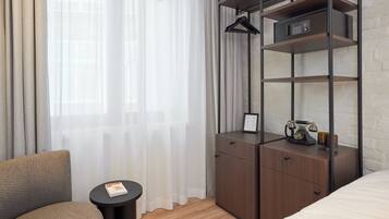 A hotel room with a bed, a wooden nightstand, a small table, a chair, and a wardrobe with shelves.