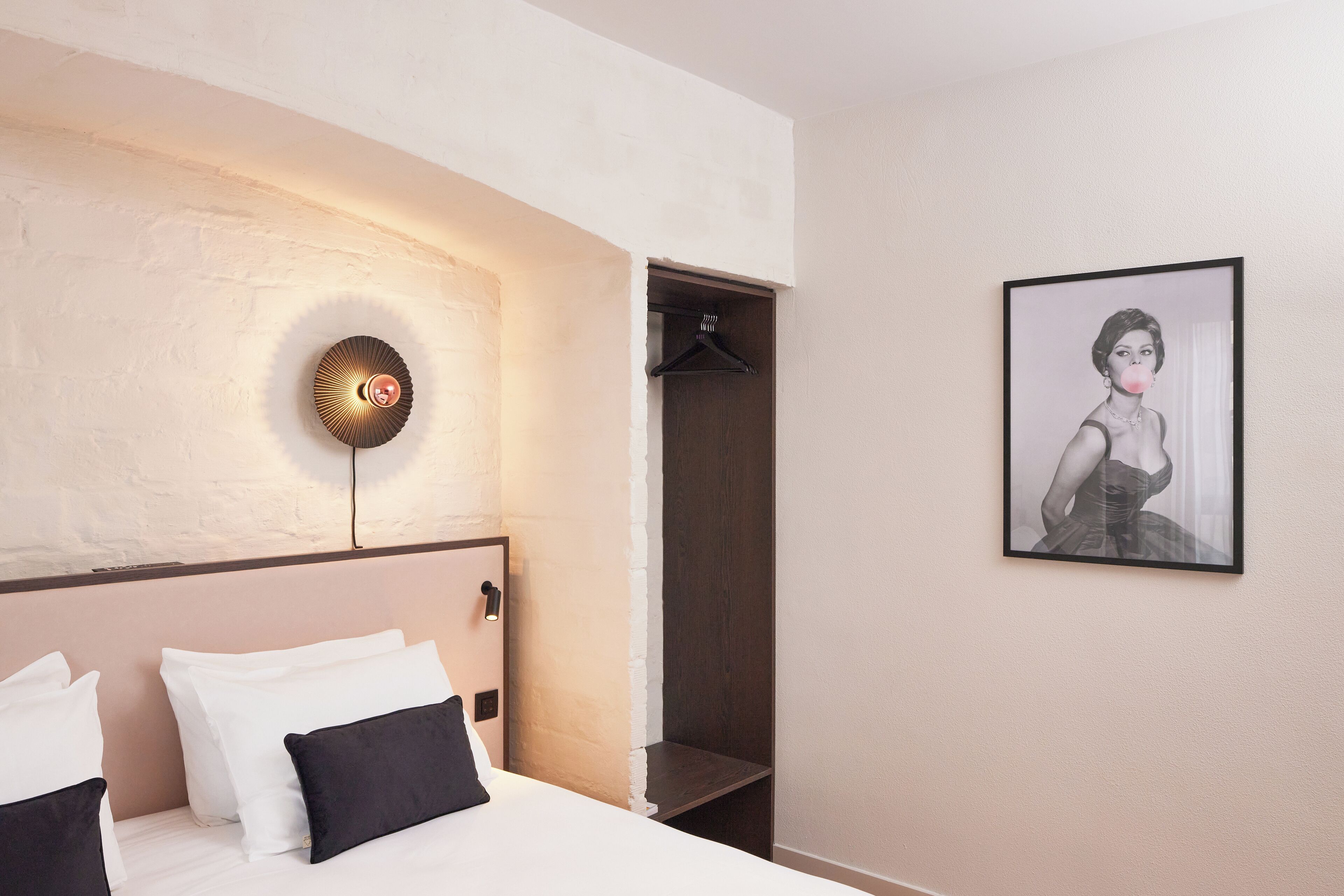 A hotel room with a bed, a coat rack, a framed picture on the wall, and a wall-mounted light fixture.