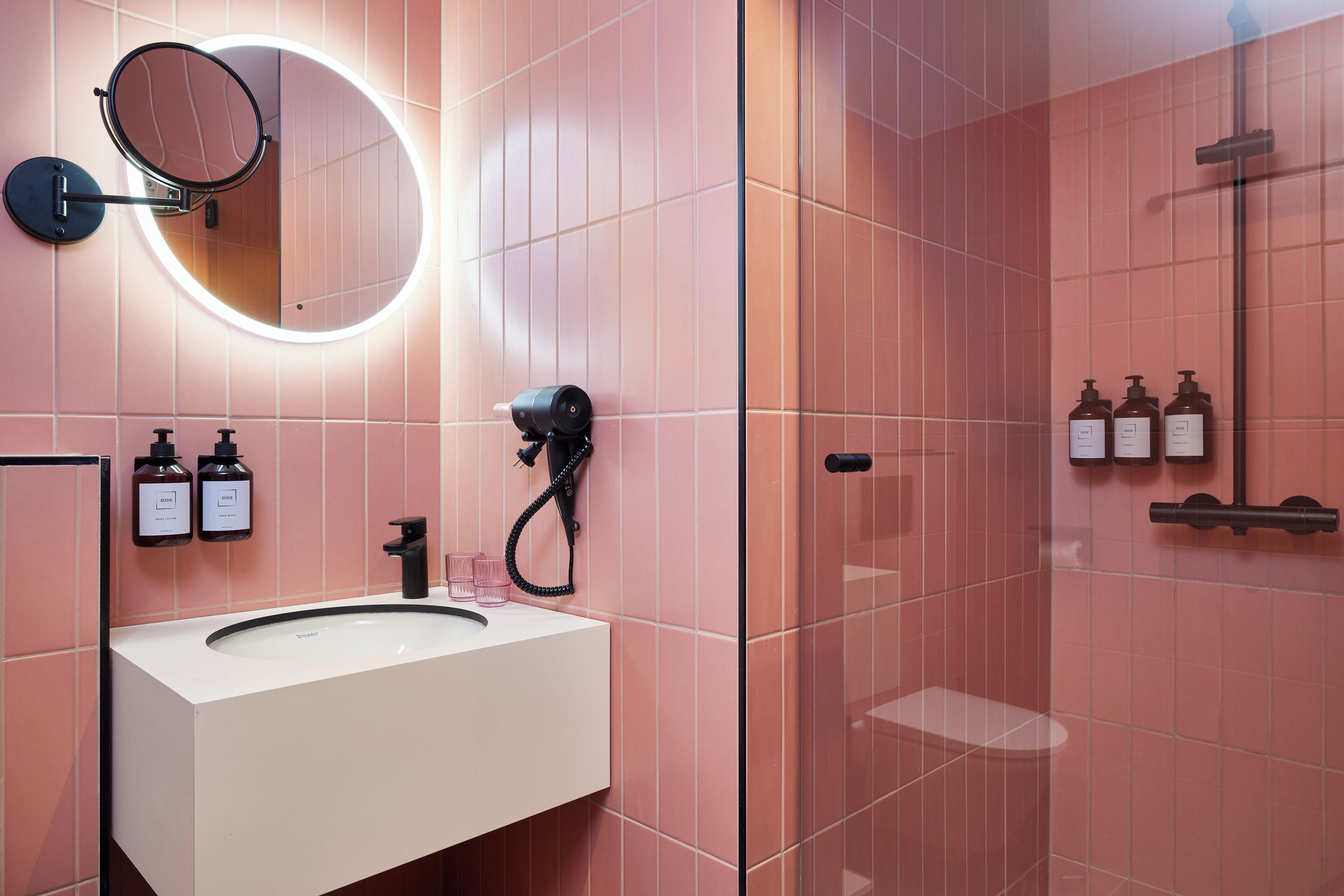 A bathroom with a white sink, a round mirror with a light, and three bottles of toiletries on the wall.