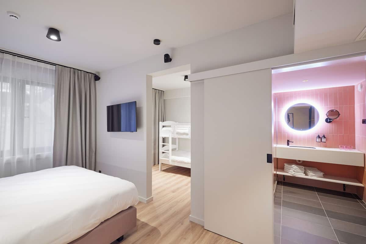 A modern hotel room with a bed, a bathroom with a pink vanity, and a view of the hallway.