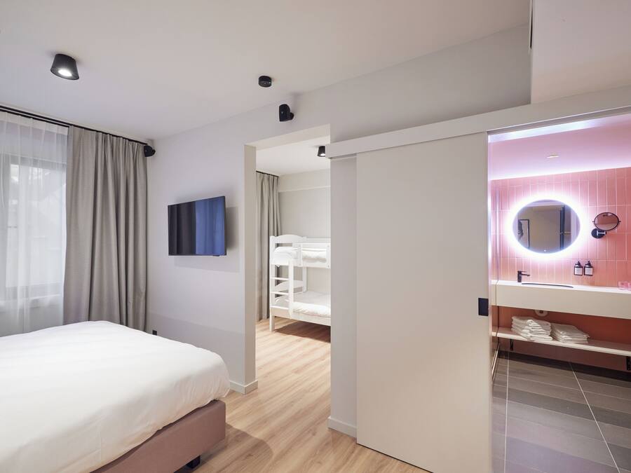 Une chambre d’hôtel moderne, avec un lit, une salle de bain dotée d’un meuble-lavabo rose et donnant sur le couloir.