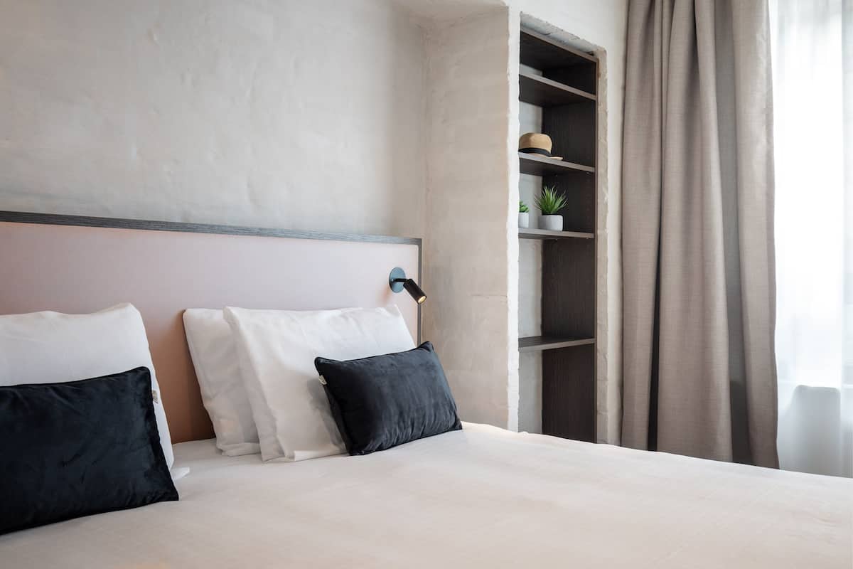 A neatly made bed with white and black pillows, a bedside lamp, and a shelf with a plant and hat.