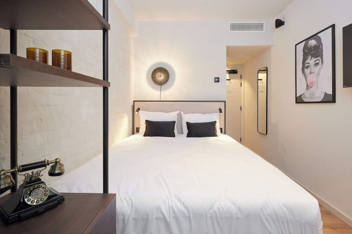 A hotel room with a bed, a telephone, a shelf with items, and a wall-mounted light.