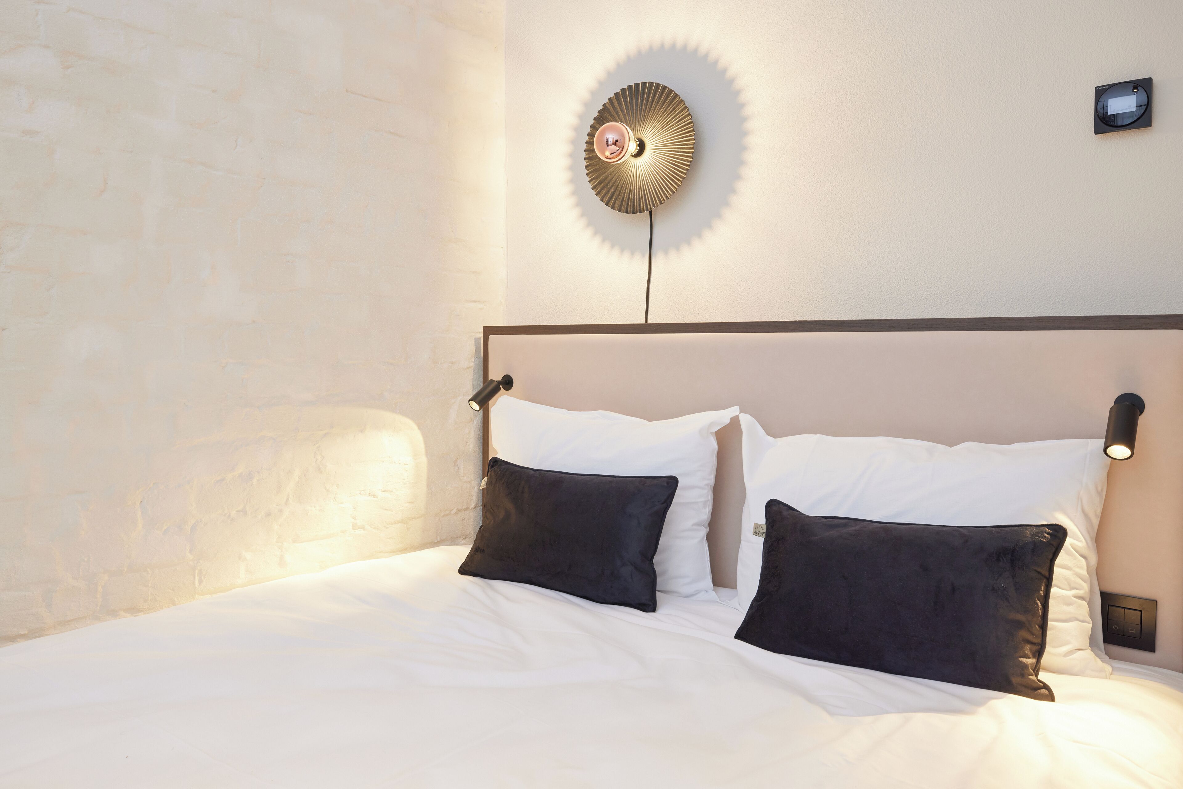A neatly made bed with white linens and two black pillows, a wall-mounted light fixture, and a textured wall.