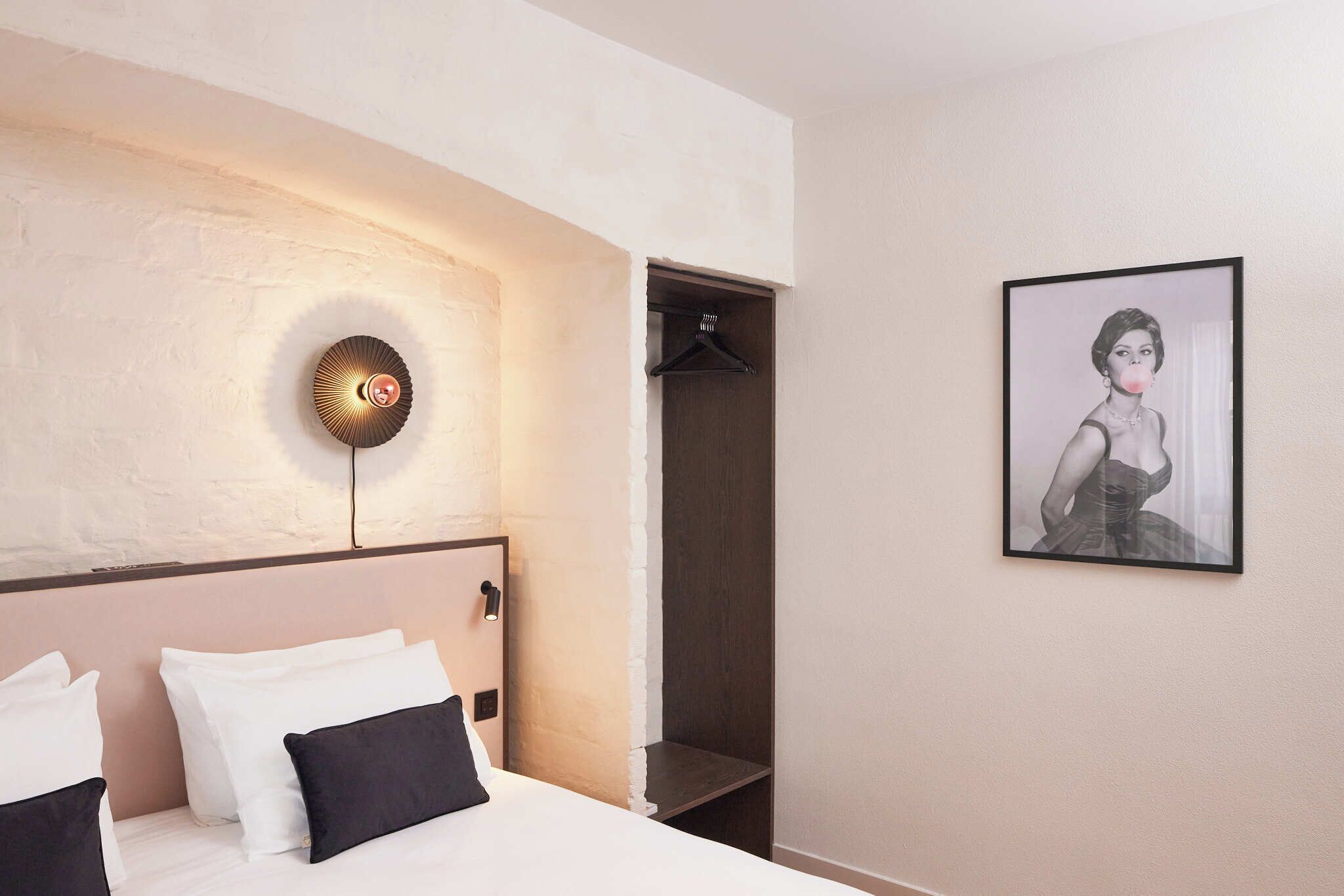 A hotel room with a bed, a wall-mounted light fixture, a coat rack, and a framed picture on the wall.