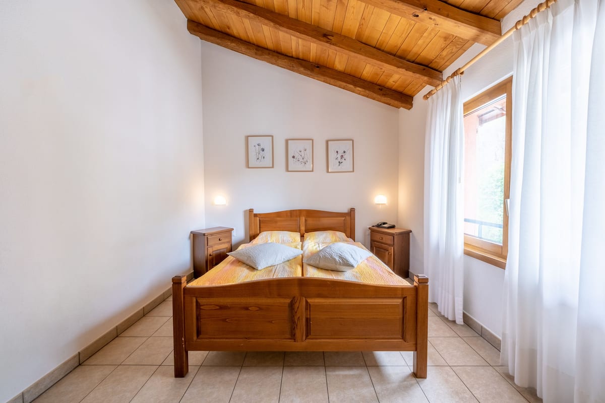 Uma cama de madeira com duas almofadas, mesas de cabeceira e luminárias fixadas na parede num quarto com teto de madeira e janela com cortinas.
