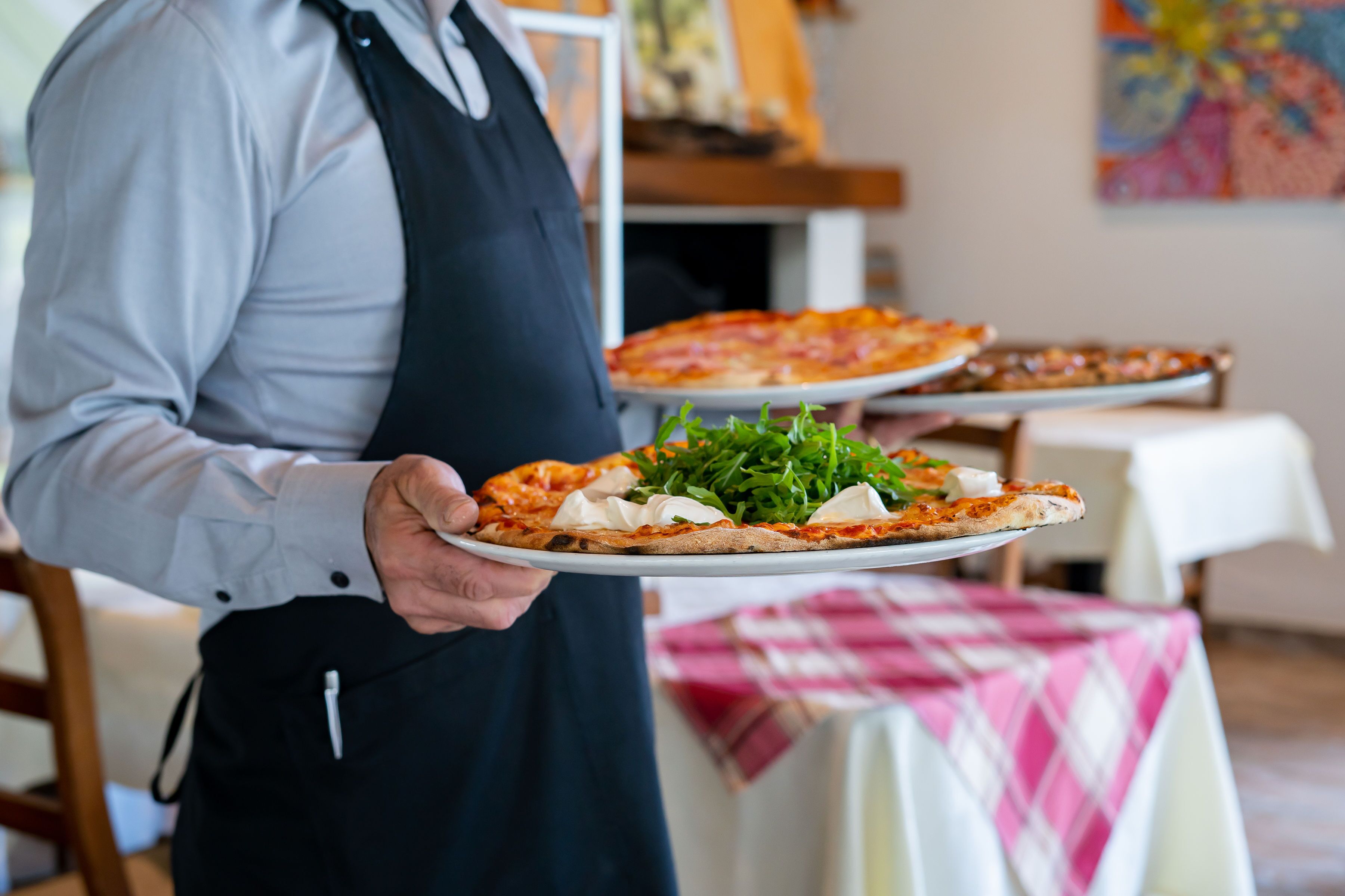 Une personne tenant une pizza sur une assiette blanche, avec une autre pizza sur un plateau en arrière-plan.