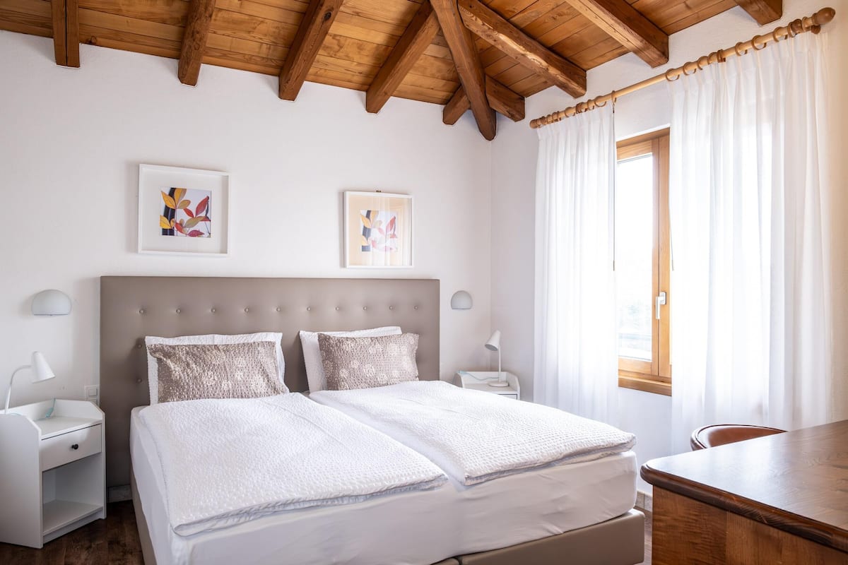 Quarto com cama, mesas de cabeceira, escrivaninha e janela com cortinas.