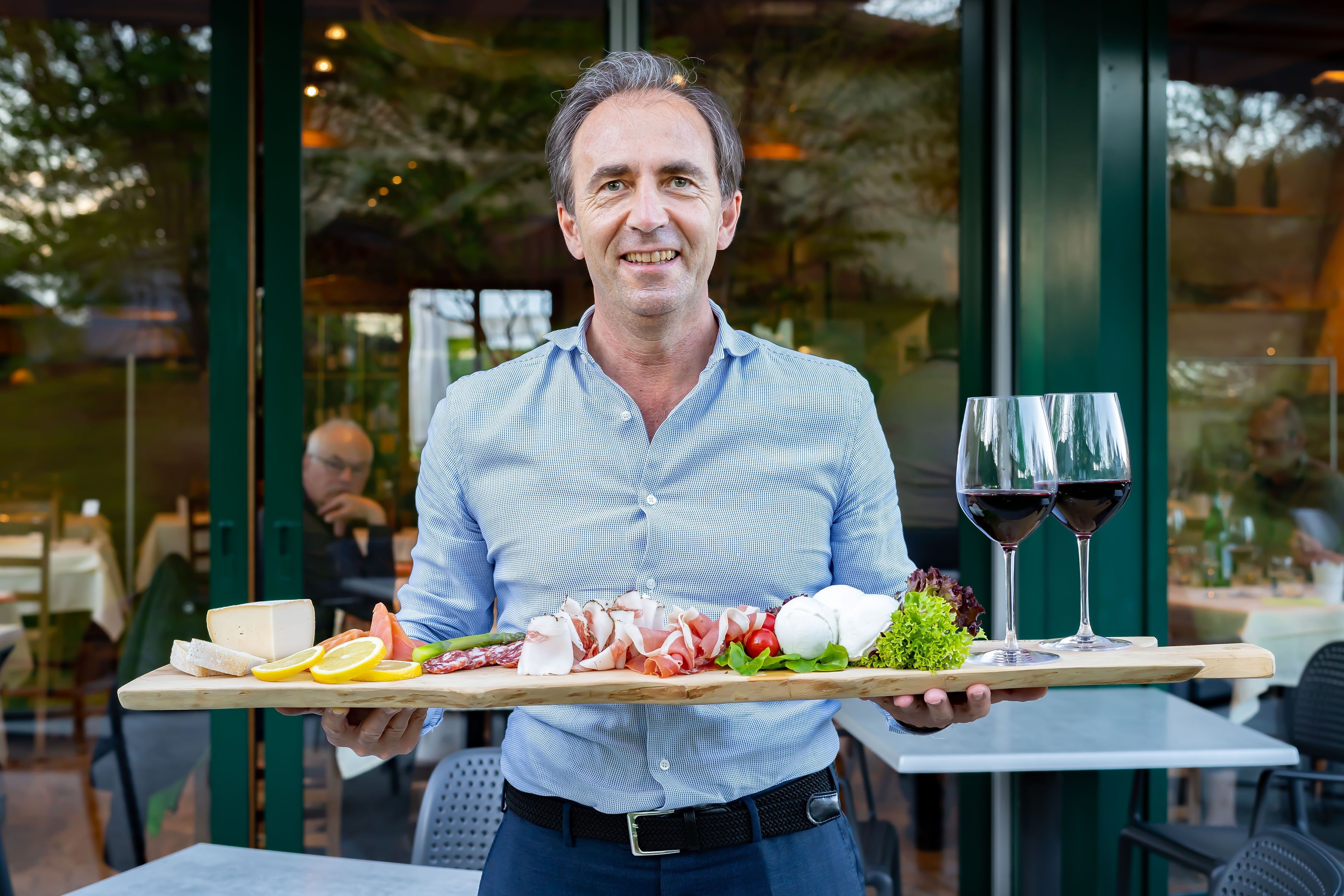 Una persona sosteniendo una bandeja con queso, carnes, vegetales y dos copas de vino tinto.