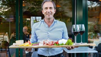 Una persona sosteniendo una bandeja con queso, carnes, vegetales y dos copas de vino tinto.