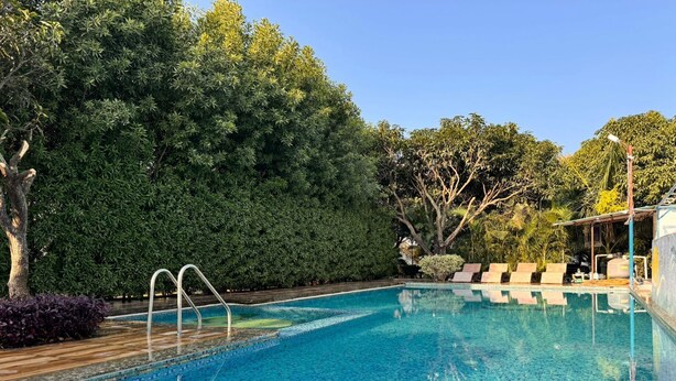 A clear swimming pool with a tiled edge, surrounded by lush greenery and a wooden deck. - Seven Corbett Resort (Kaladhungi)