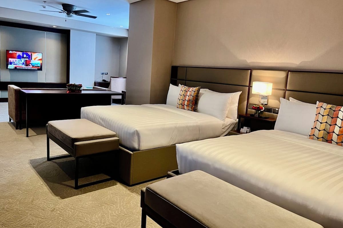 A hotel room with two beds, a desk, a TV, and a ceiling fan.