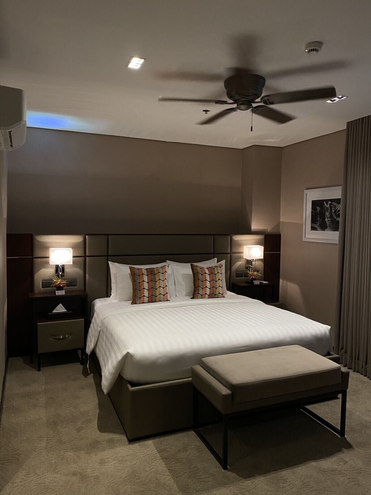 A hotel room with a large bed, two bedside tables with lamps, a ceiling fan, and a window with curtains.