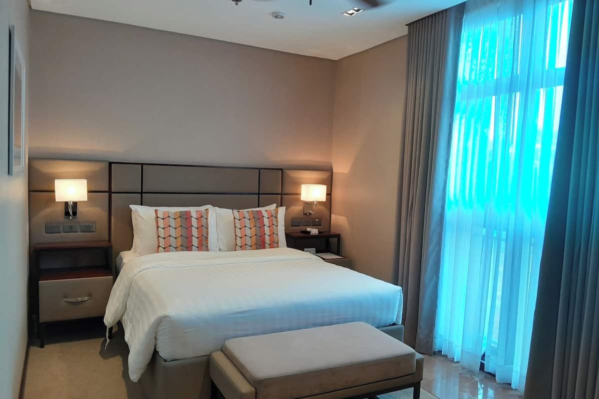 A modern hotel room with a large bed, bedside lamps, a nightstand, a bench, and a view of the outdoors through sheer curtains.