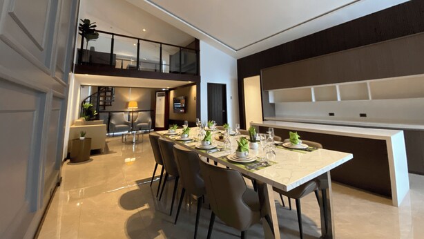 A modern dining area with a long table, multiple chairs, and a kitchen area in the background.