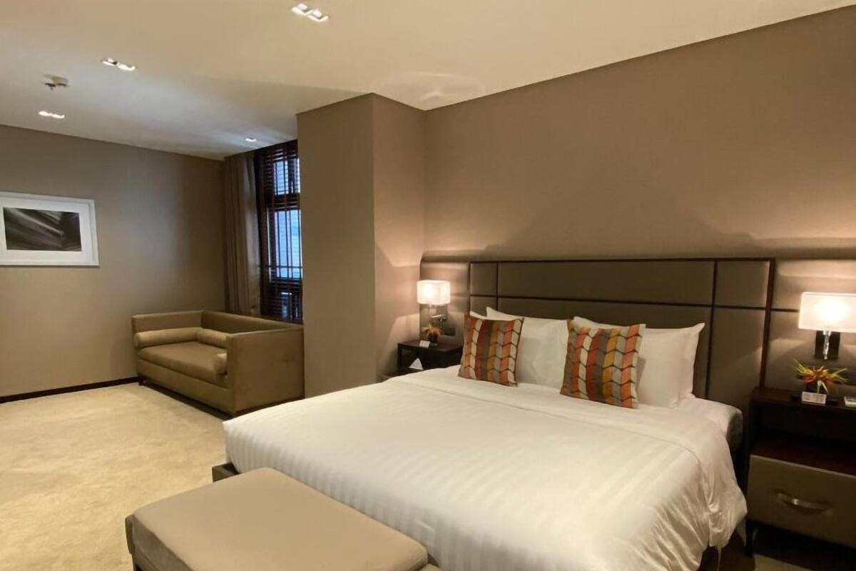 A hotel room with a large bed, a sofa, a nightstand, and a window with blinds.