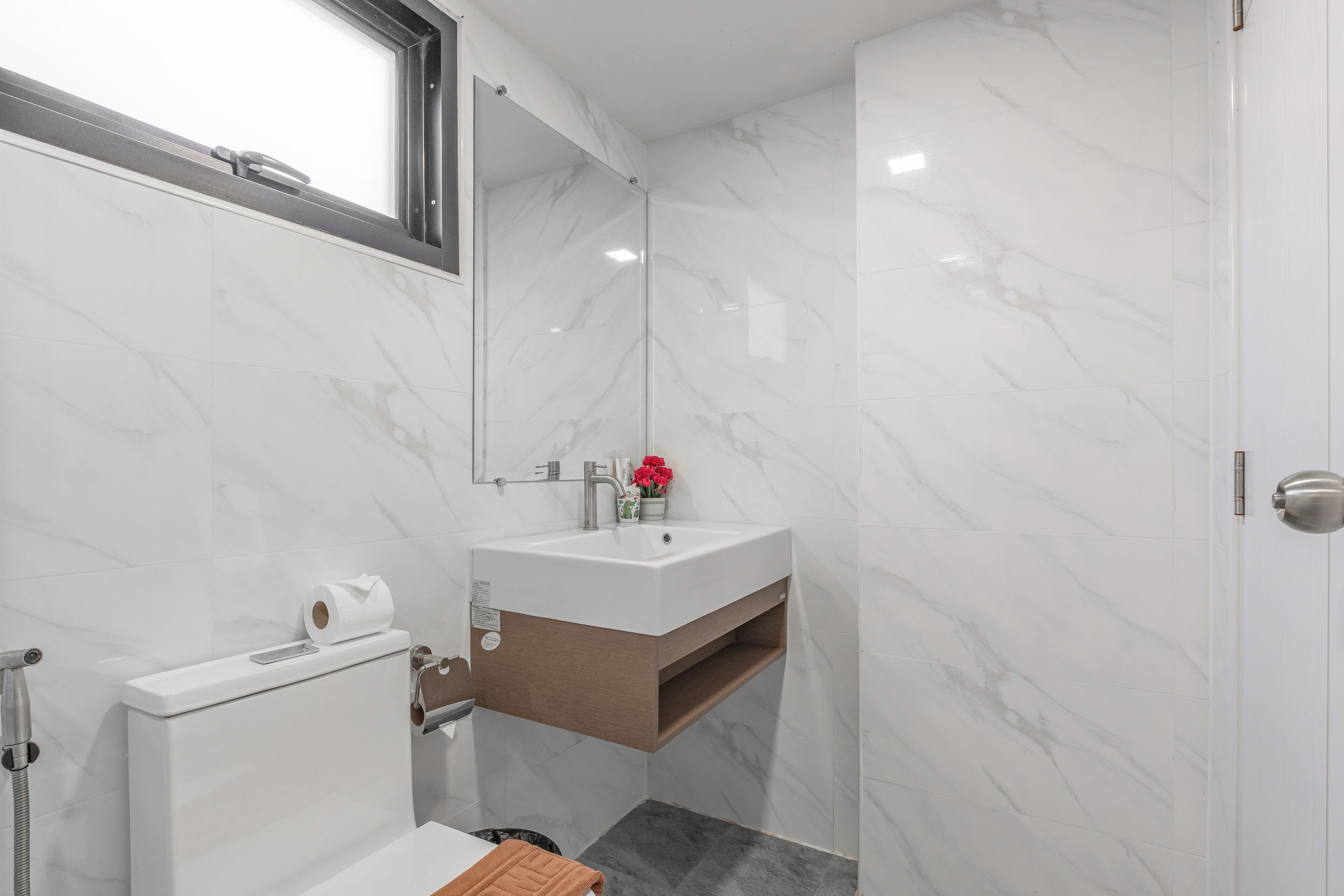 A bathroom with a white marble wall, a white sink with a mirror above it, and a toilet with a roll of toilet paper.
