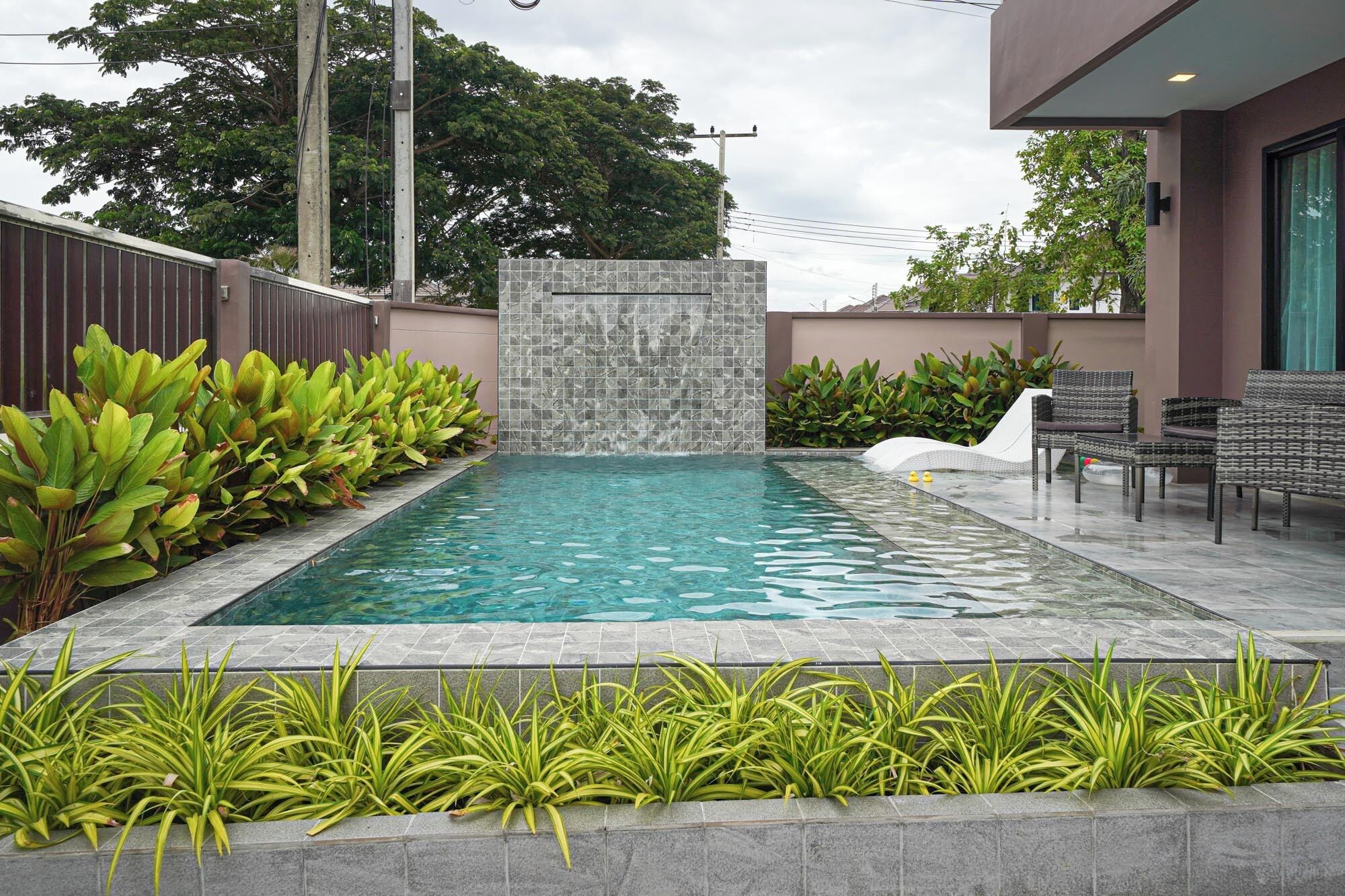 A rectangular swimming pool with a stone waterfall feature, surrounded by lush green plants and a modern outdoor seating area.