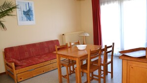 A small, cozy room with a red sofa, wooden dining table, and chairs. There's a radiator, a lamp, and a window with curtains. - Beautiful T3 apartment with balcony and magnificent view (Font-Romeu-Odeillo-Via)