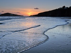 A beach with waves gently lapping at the shore, a sunset in the background, and a silhouette of a hill with buildings. - 7 Beachdown (Kingsbridge)