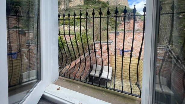 Vista desde una ventana con una valla de hierro forjado, un muro de ladrillo y un castillo al fondo.