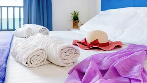 Un lit bien fait, avec des serviettes blanches pliées en rouleaux, une couverture violette et un chapeau rouge et beige.