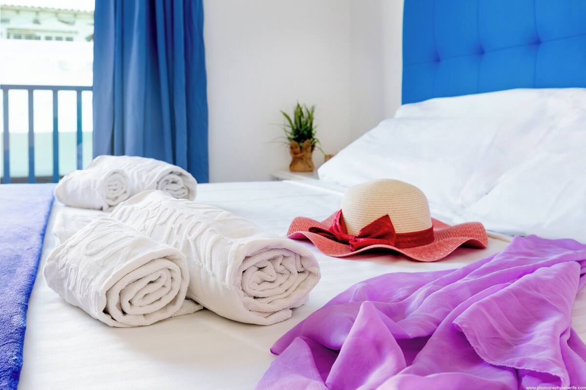 Een net opgemaakt bed met opgerolde witte handdoeken, een paarse deken en een rode en beige hoed.