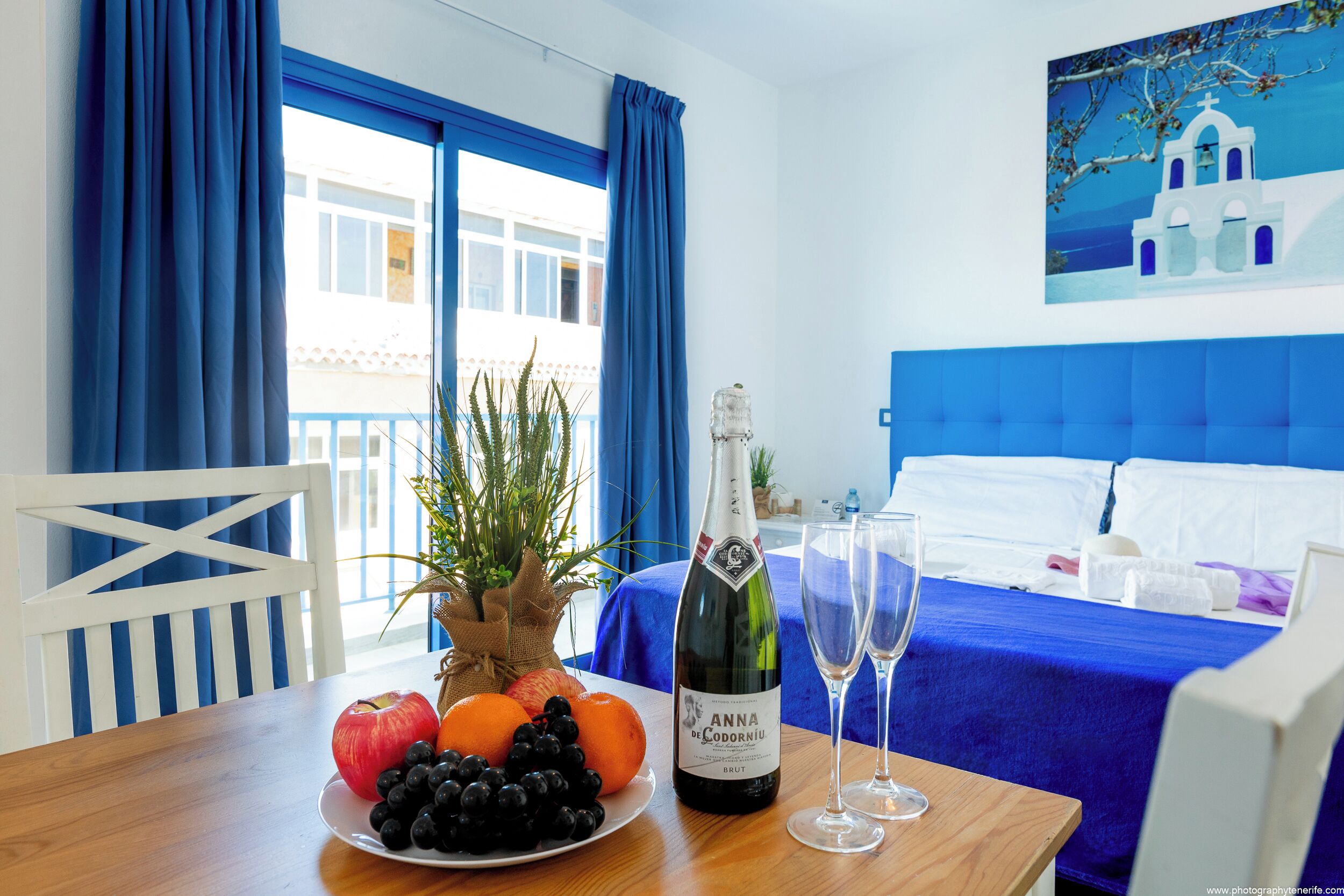 A hotel room with a bed, a dining table with fruits and a bottle of champagne, and a view of a building through the window.