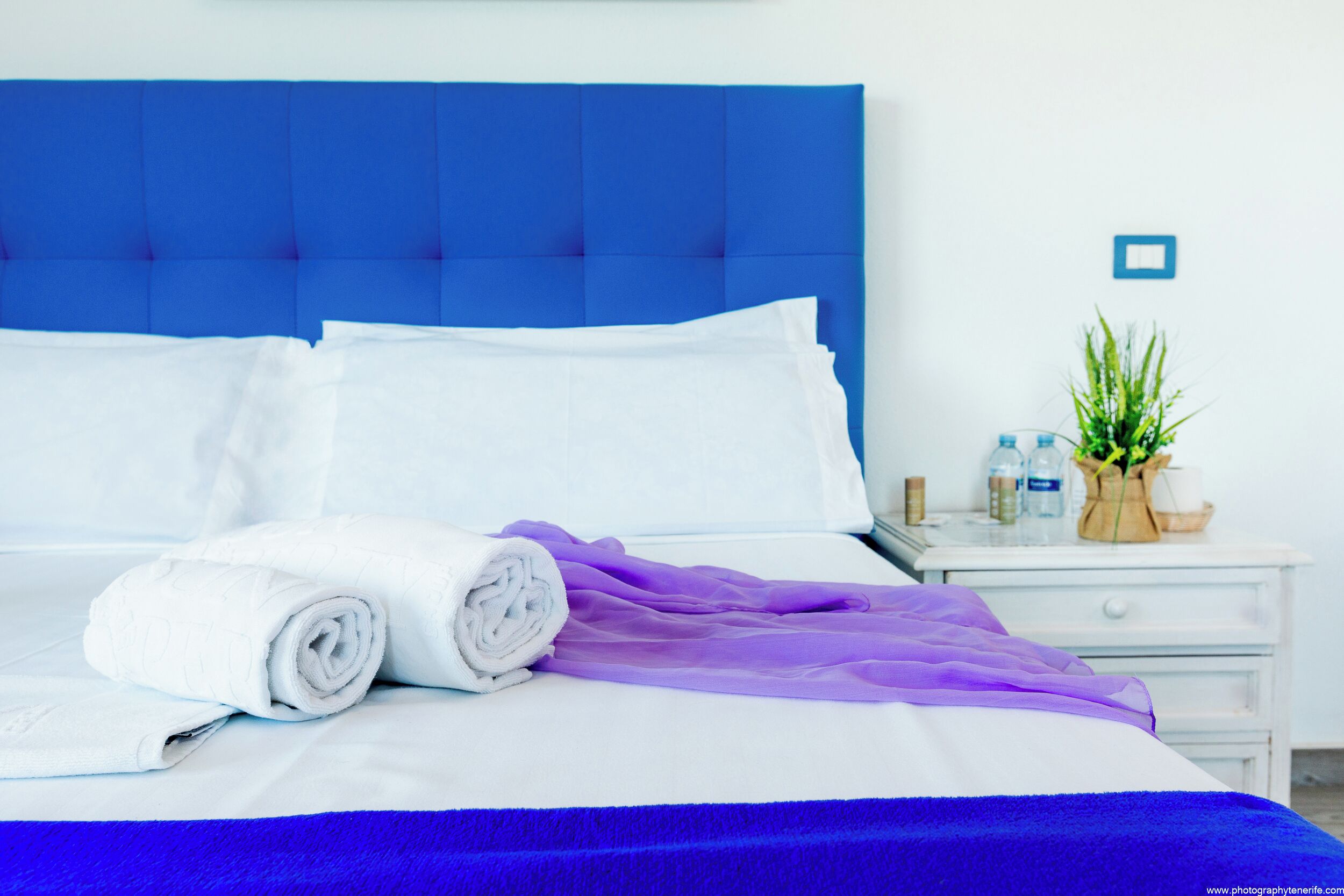 Una cama bien tendida con ropa de cama blanca y morada, un cabecero azul y una mesita de noche con una planta y artículos de aseo.