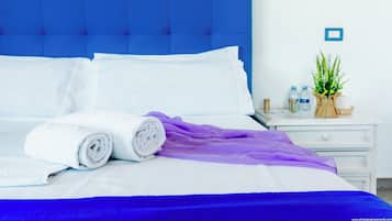Una cama bien tendida con ropa de cama blanca y morada, un cabecero azul y una mesita de noche con una planta y artículos de aseo.