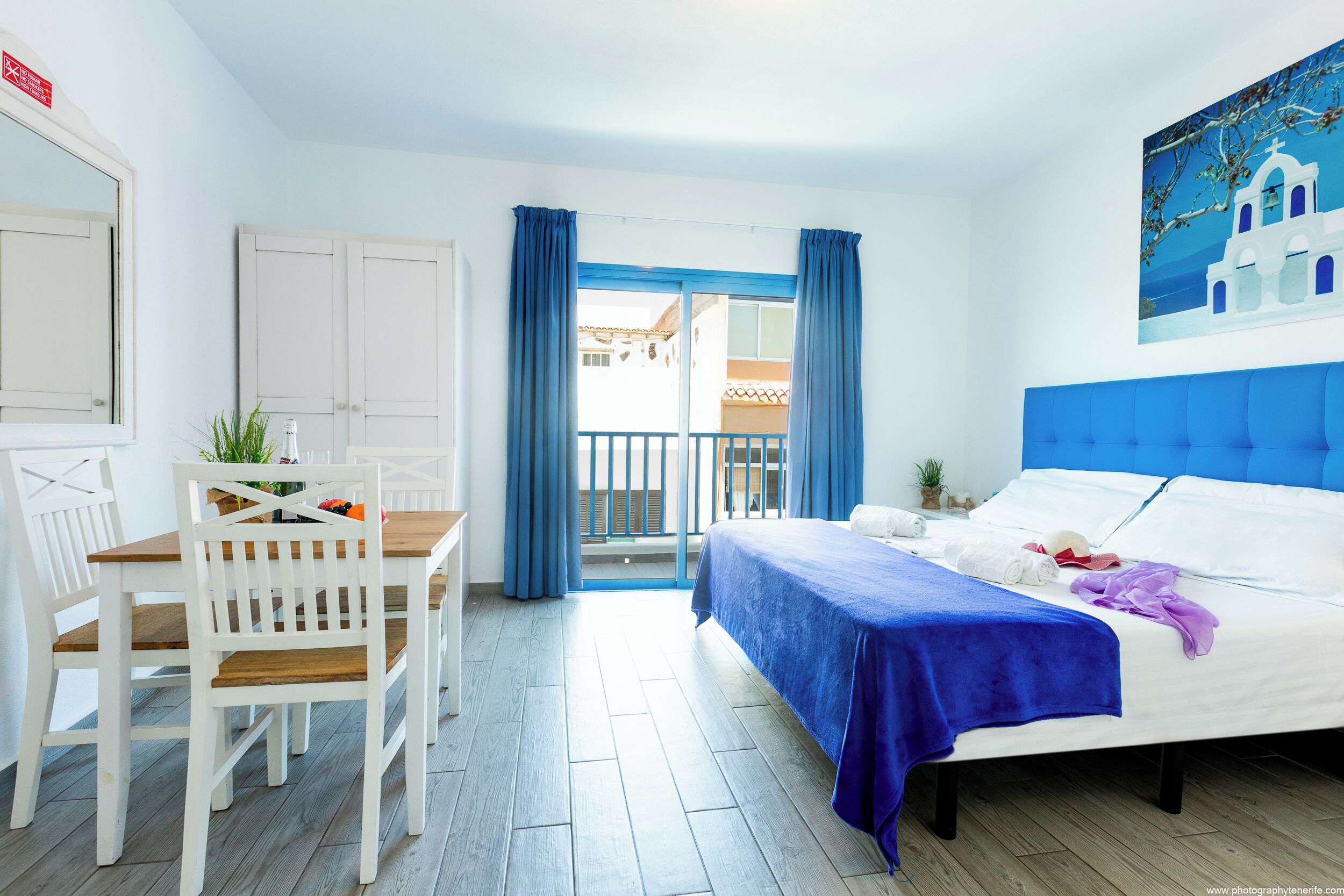 A hotel room with a bed, a desk with chairs, a balcony with blue curtains, and a painting of a white building.