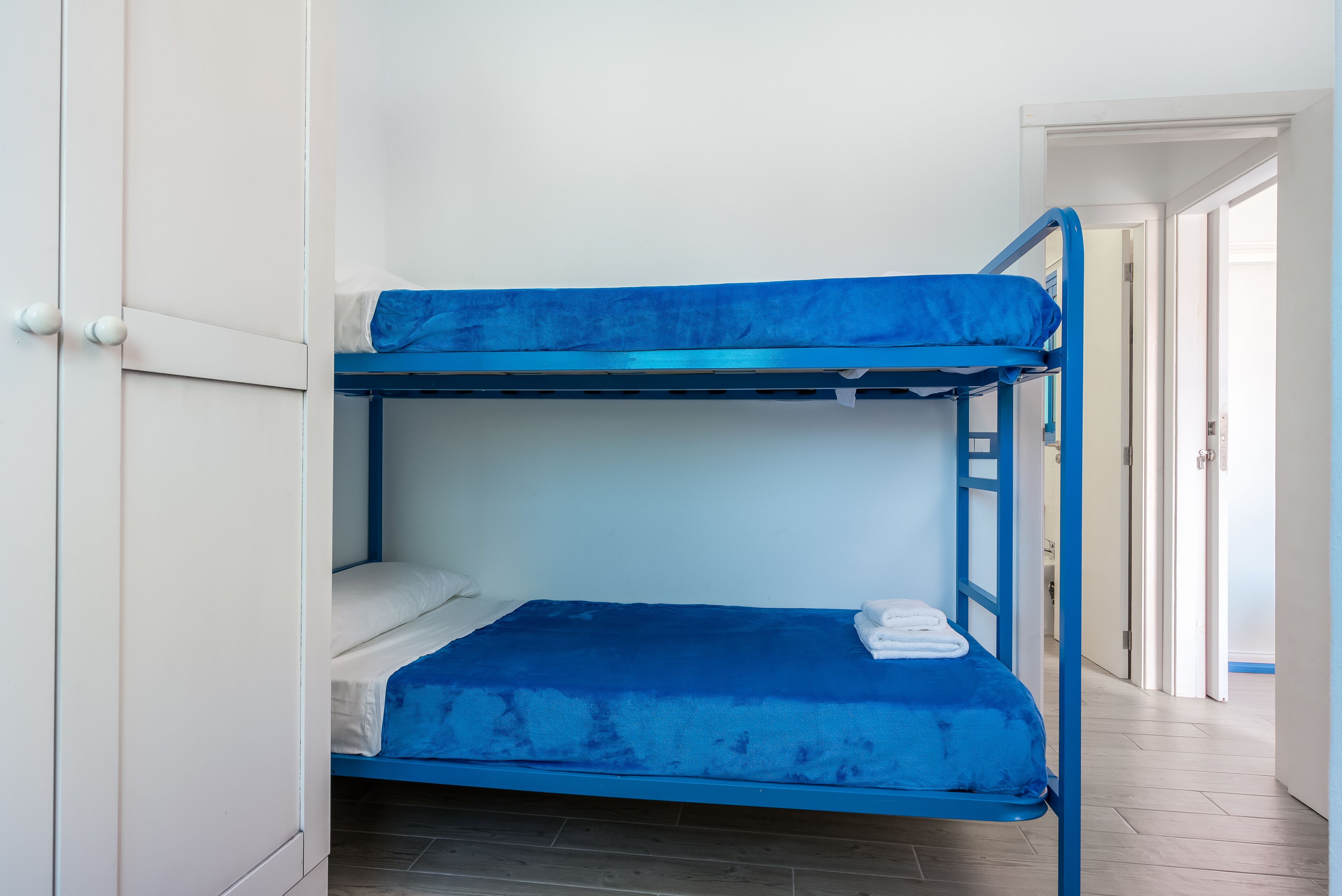 Un letto a castello con materasso blu e cuscini bianchi, un'armadio di colore bianco e un pavimento in legno.